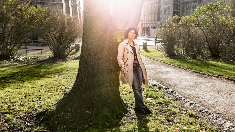 Kvinna står lutad mot ett stort träd med solen som strålar mot dig