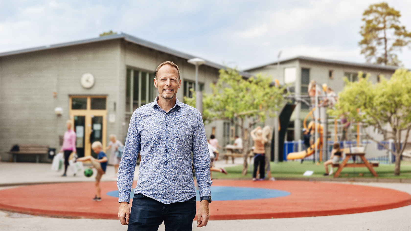 Örjan Johansson står framför en skola med lekande barn
